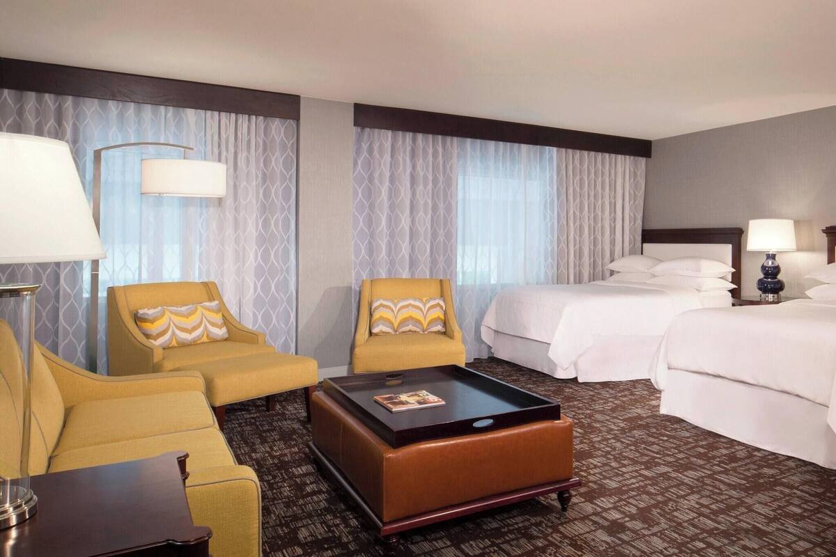 A hotel room with two yellow armchairs, a brown ottoman, a large bed, two bedside tables with lamps, and a patterned carpet.