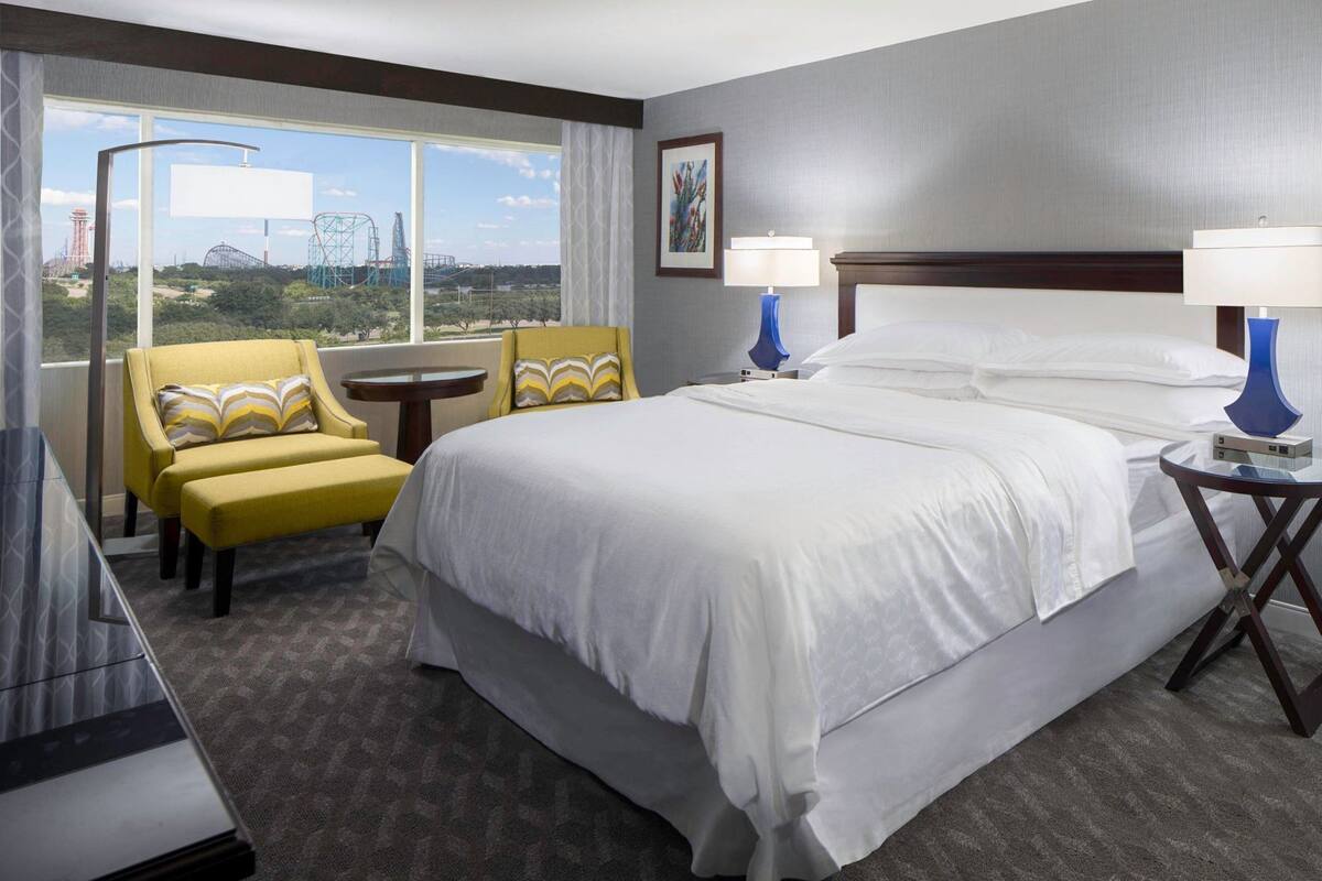 A hotel room with a large bed, two bedside lamps, a chair, a small table, and a view of amusement park rides.