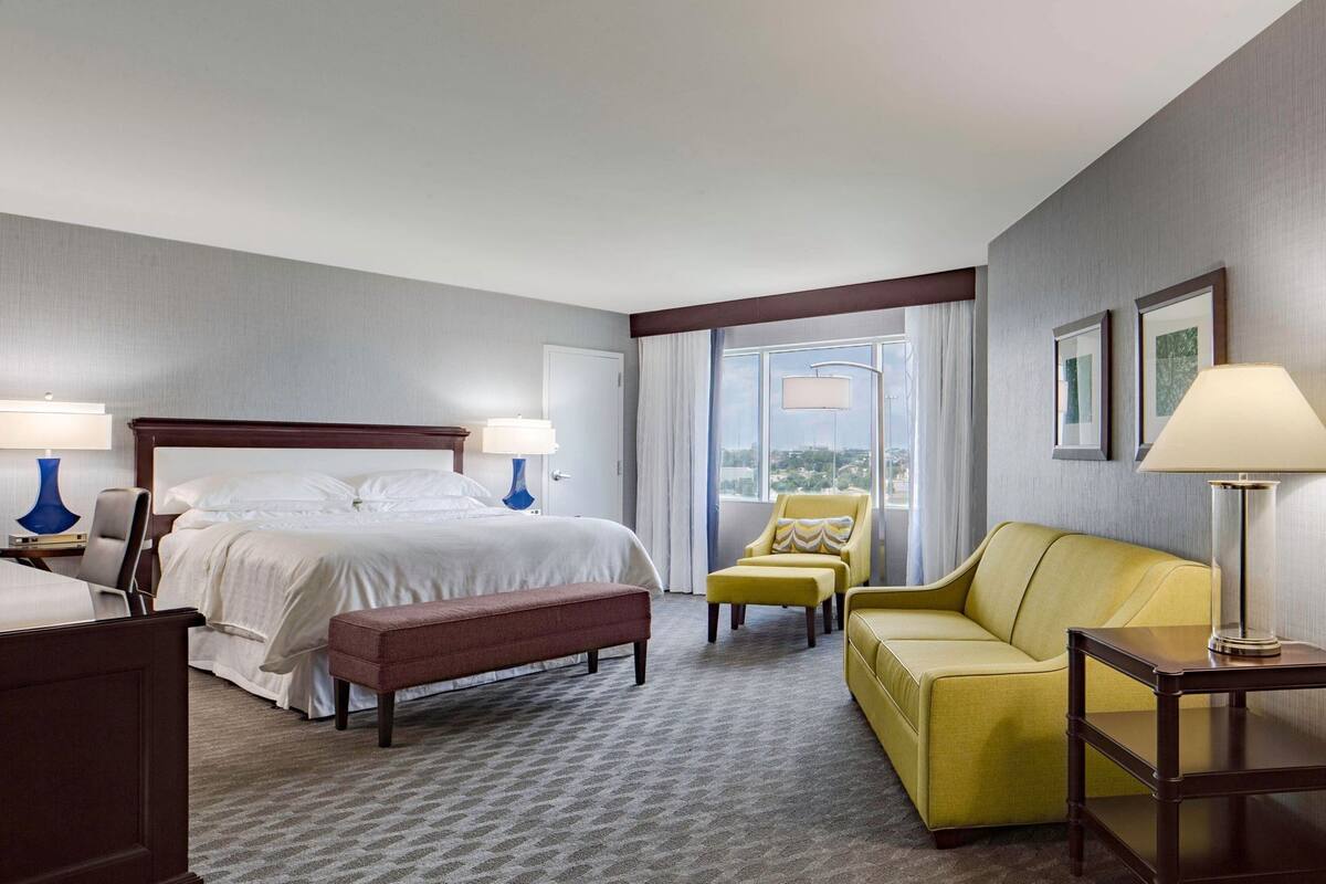 A hotel room with a bed, a yellow armchair, a bench, a sofa, and a lamp.