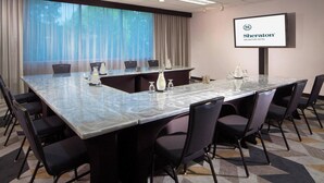 A conference room with a large table, chairs, a projector screen displaying the Sheraton logo, and a large window with curtains. - Sheraton Arlington (Arlington)