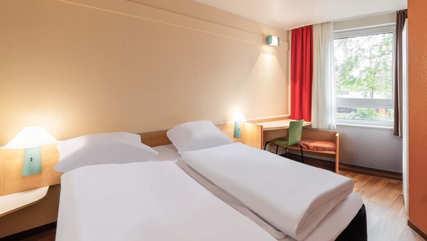A hotel room with two beds, a desk, a chair, and a window with curtains.