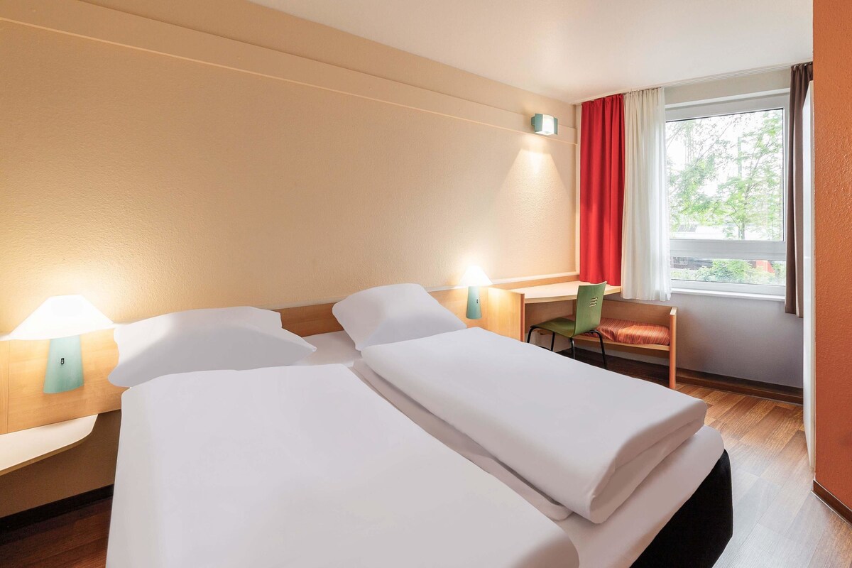 Une chambre d’hôtel avec deux lits, un bureau, une chaise et une fenêtre avec des rideaux.