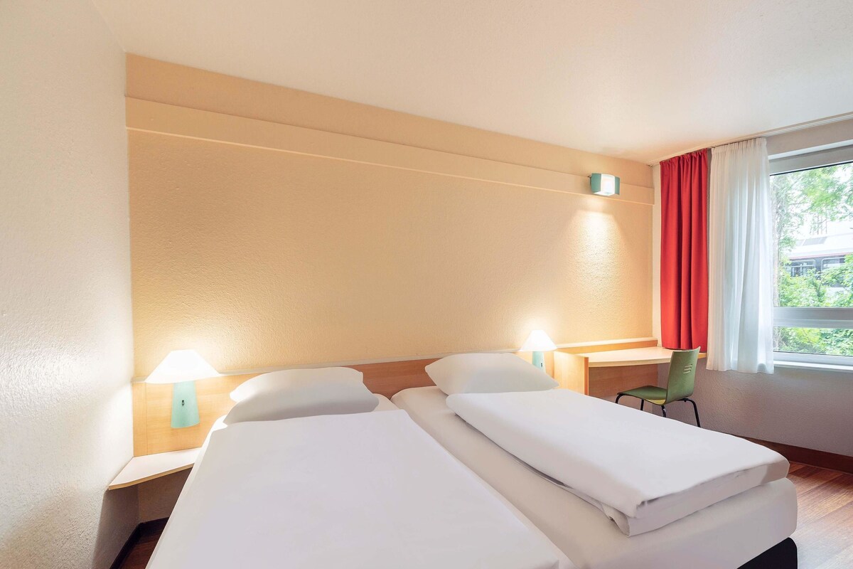 Une chambre d’hôtel avec deux lits, un bureau, une chaise, une lampe et une fenêtre avec des rideaux.
