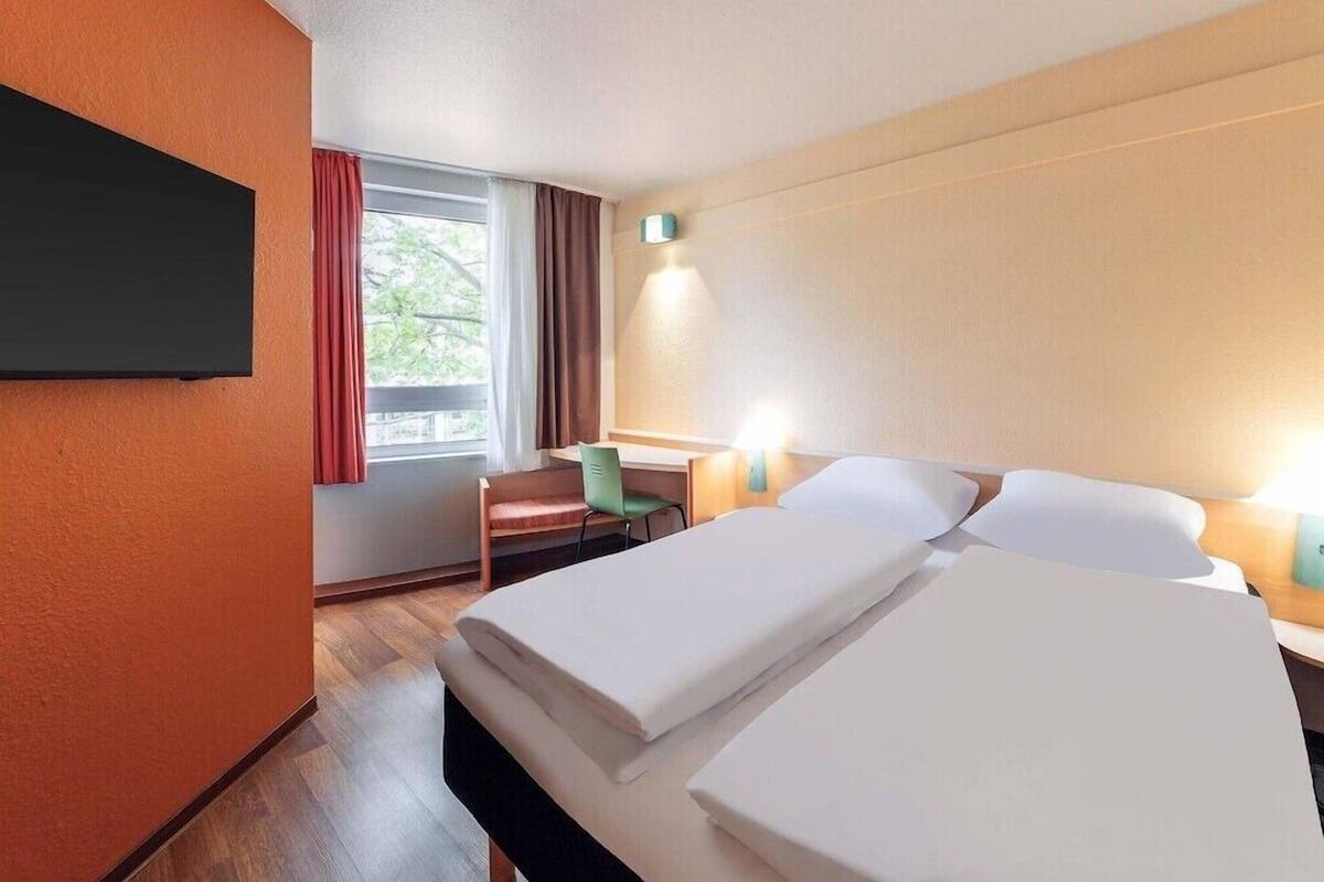 A hotel room with two beds, a desk, a chair, a TV, and a window with curtains.