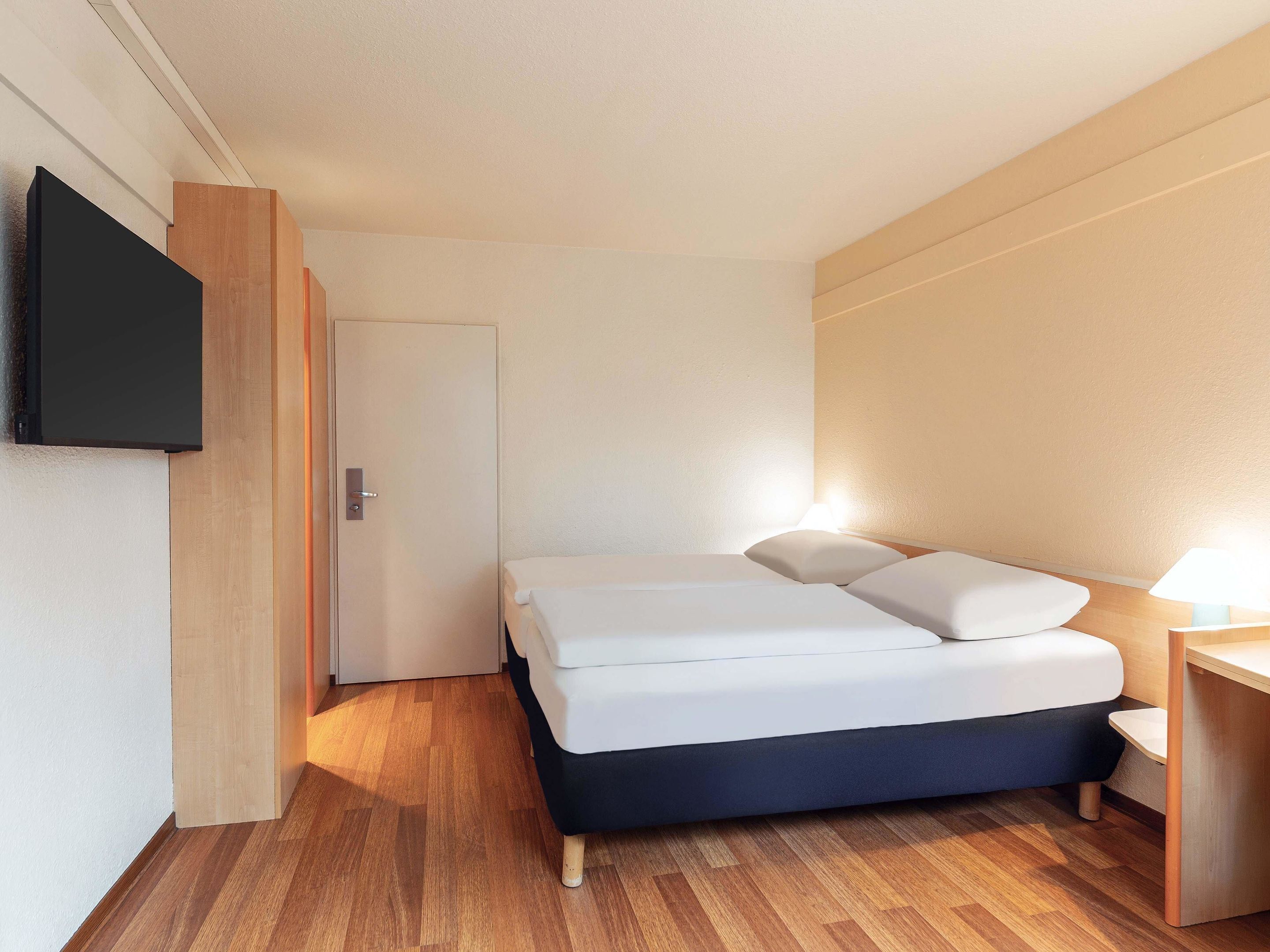 A compact hotel room with a bed, a TV mounted on the wall, and a wooden floor.