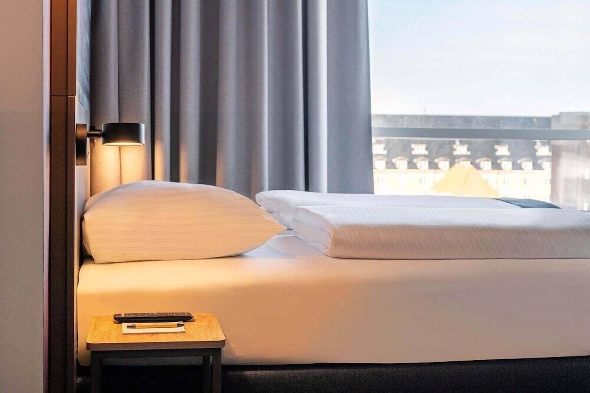 A hotel room with a bed, a nightstand, a lamp, and a view of buildings through the window.
