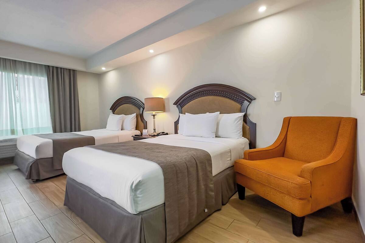 A hotel room with two beds, a chair, a bedside table, and a lamp.
