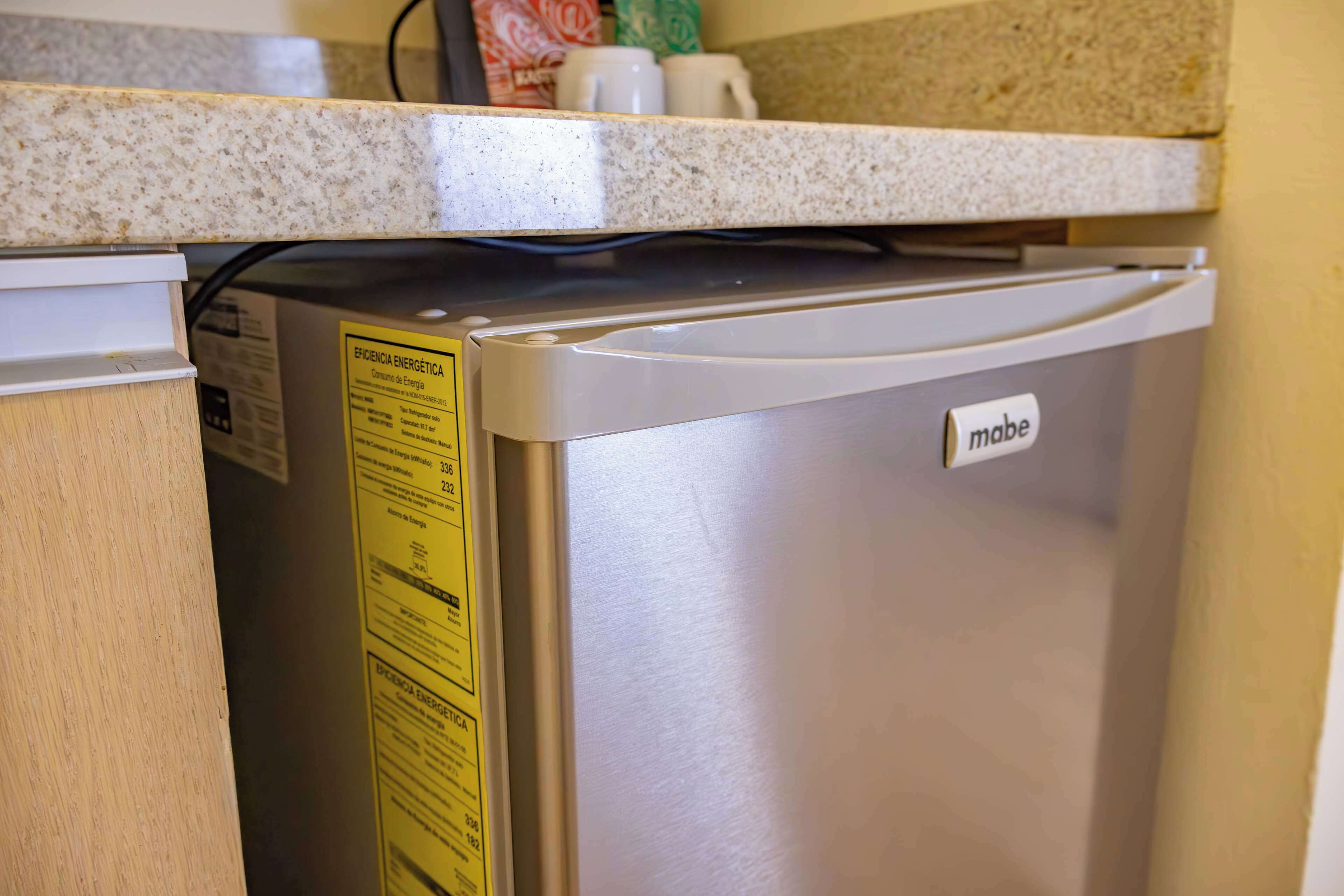 A compact, stainless steel refrigerator with a 'mabe' label, situated under a granite countertop in a kitchen setting.