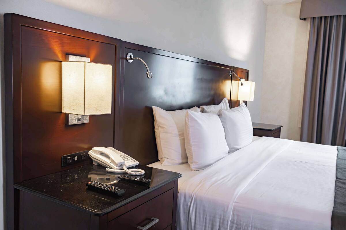 A hotel room with a wooden headboard, a bedside table with a telephone and remote control, and two wall-mounted lamps.