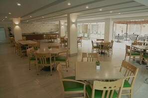 Un comedor amplio con varias mesas y sillas de madera, una cocina al fondo y ventanas grandes que ofrecen luz natural.