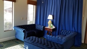 A room with a blue sofa, a lamp on a side table, and a view through a window with blinds. - The Parkwood Inn & Suites (Mountain View)