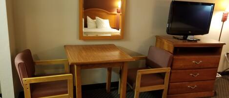A hotel room with a wooden desk, two chairs, a television, a mirror, and a dresser.