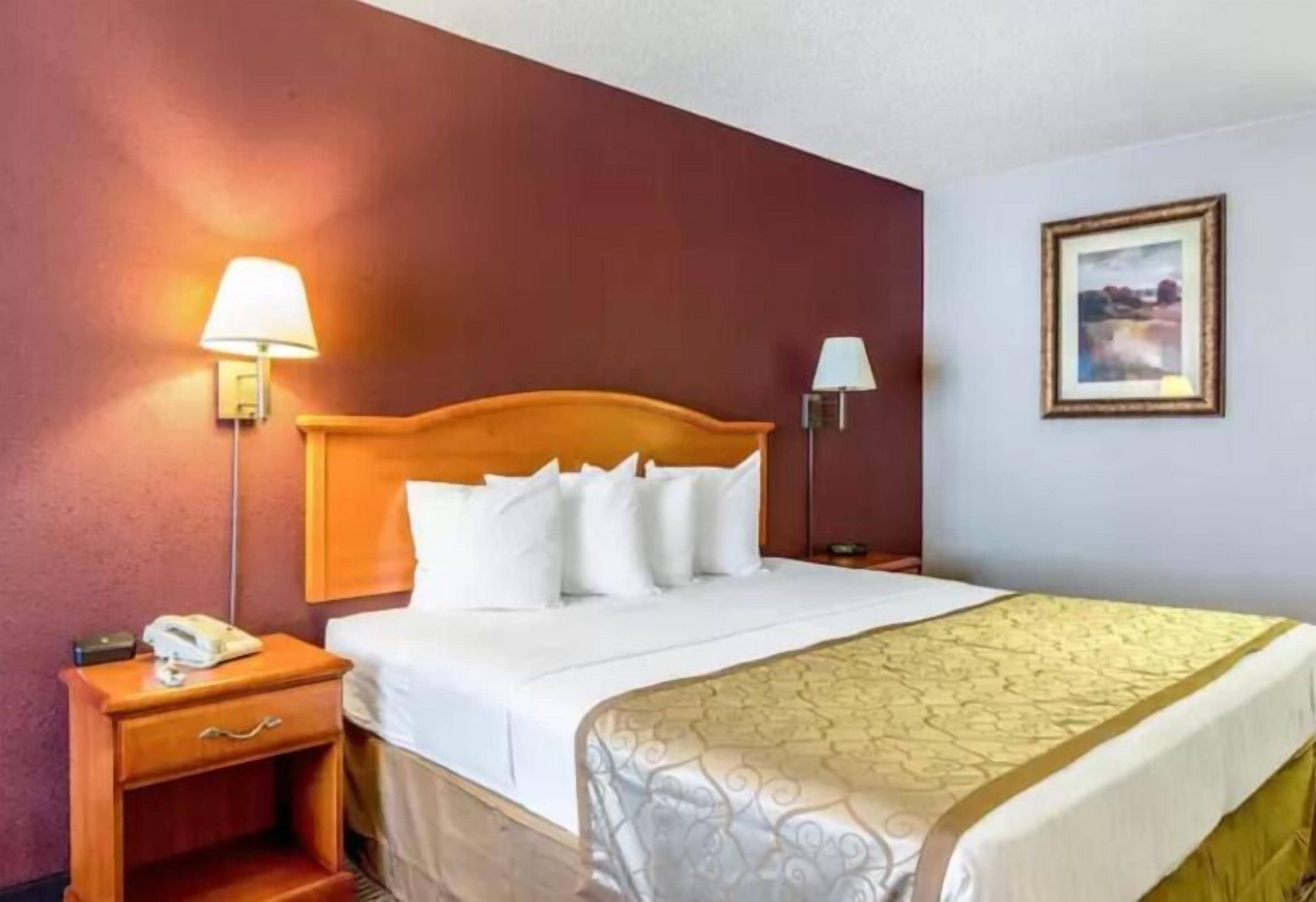 A hotel room with a wooden headboard, a white bedspread, a yellow accent bedspread, a bedside lamp, a nightstand with a phone, and a framed picture on the wall.
