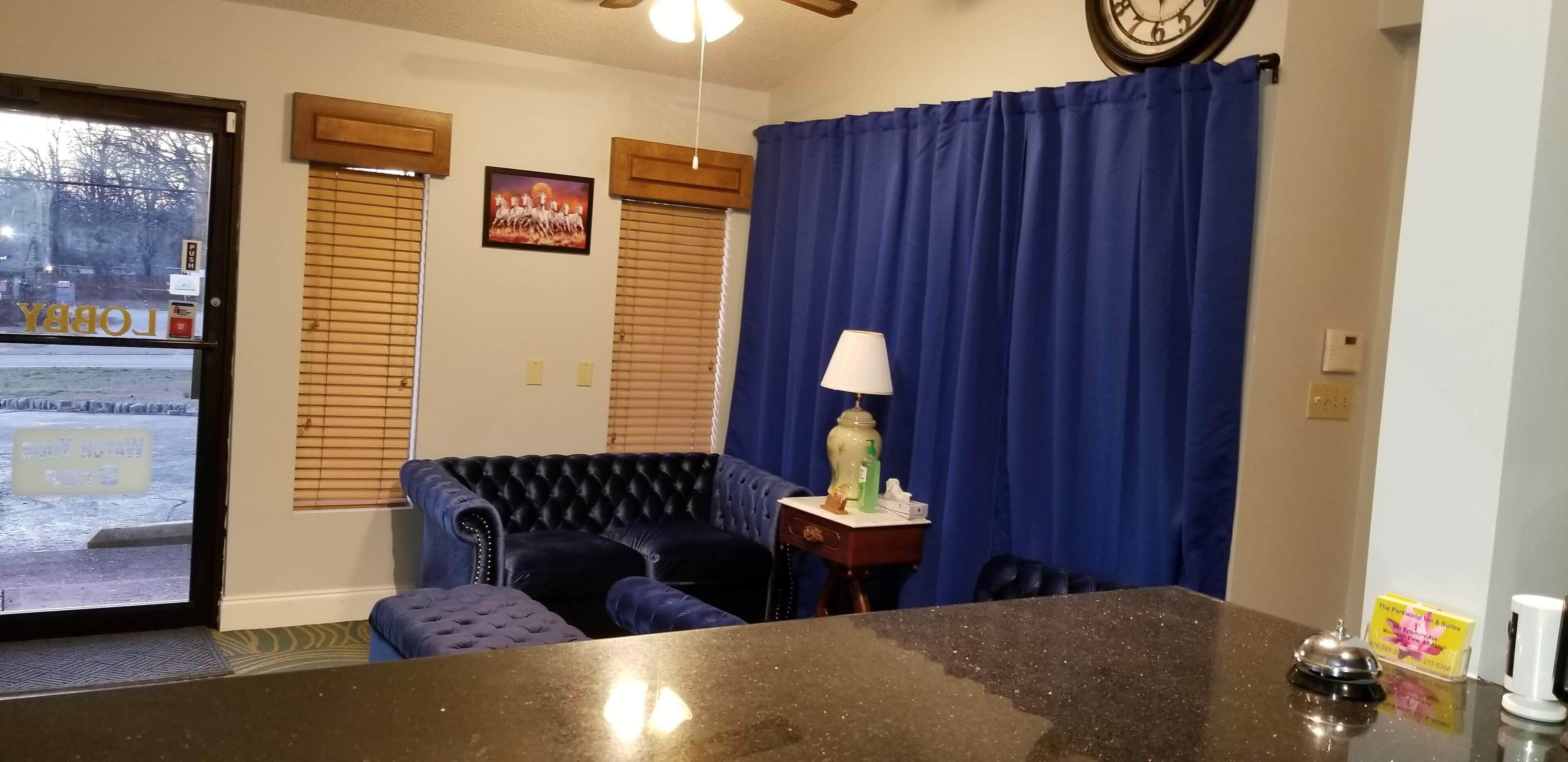 A room with a black leather sofa, a small table with a lamp, a clock on the wall, and a blue curtain.