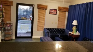 A room with a black leather sofa, a small table with a lamp, a blue curtain, and a door leading outside. - The Parkwood Inn & Suites (Mountain View)