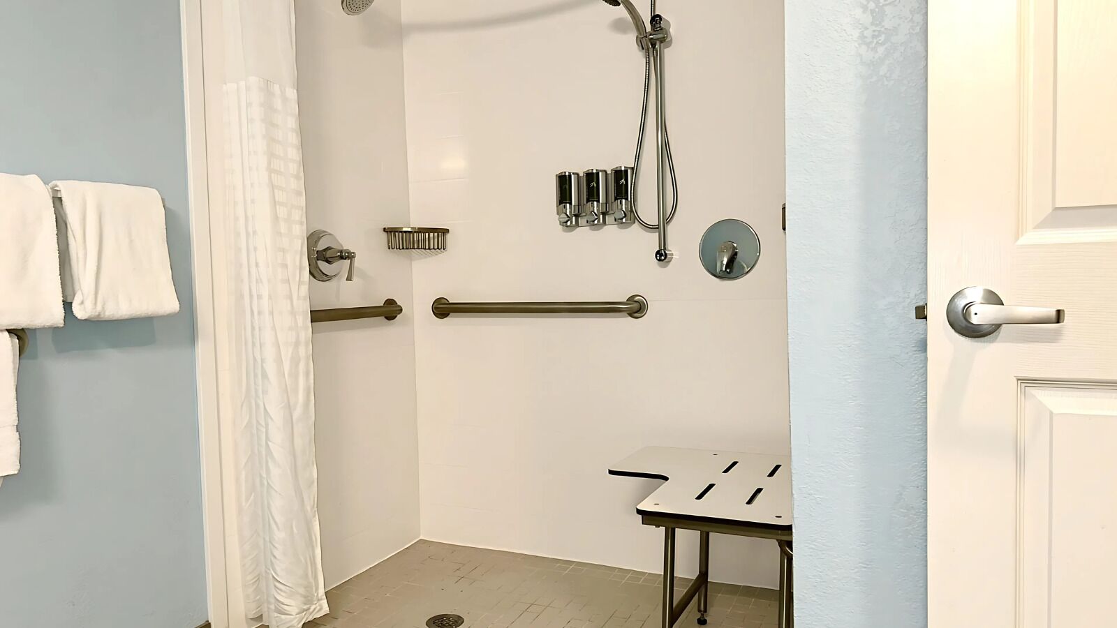 A bathroom with a shower, grab bars, a towel rack, and a small table.