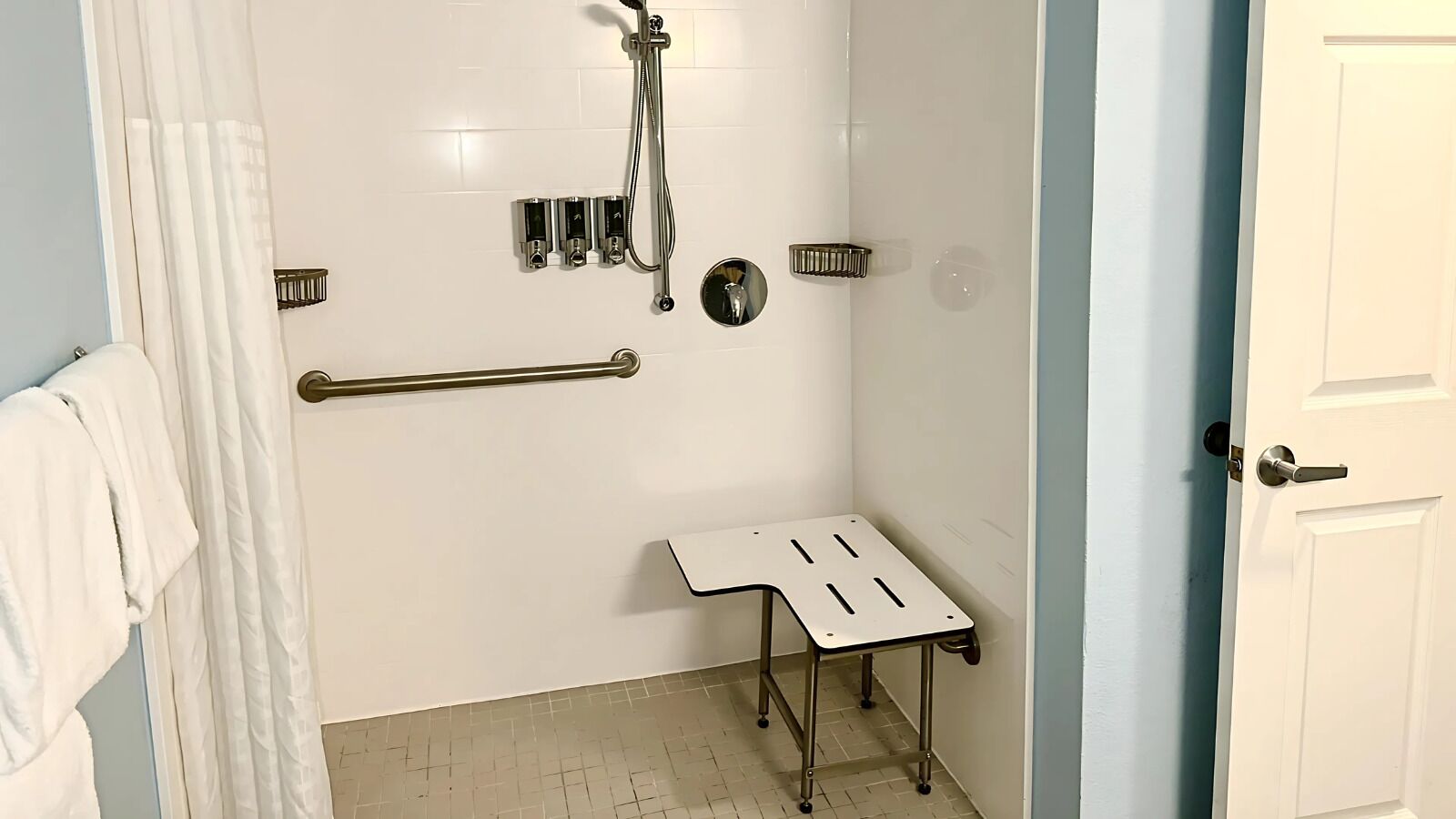 A small, enclosed shower area with a white tiled wall, a handheld showerhead, a handheld shower attachment, a small table, and a grab bar.