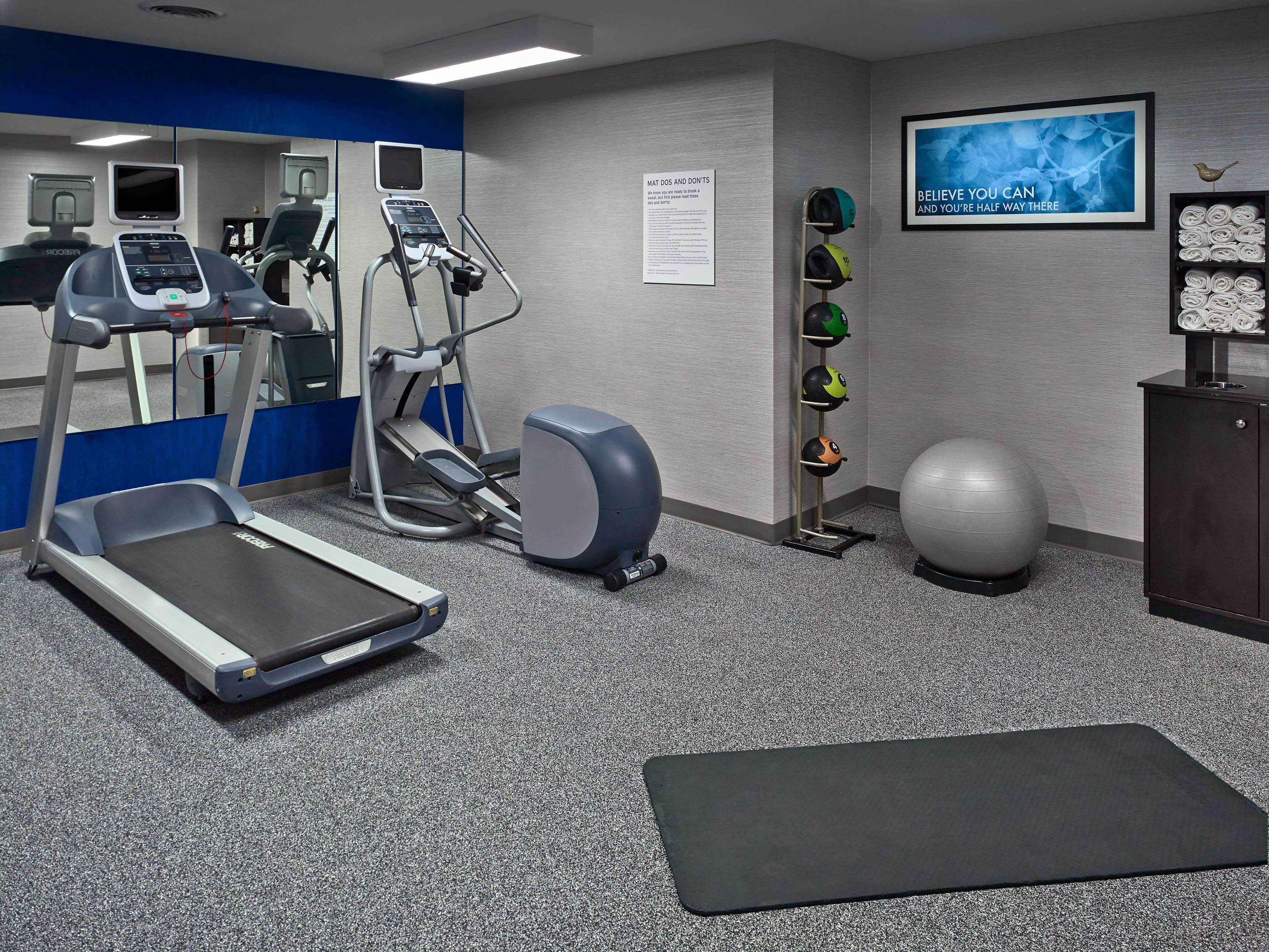 Une salle de sport équipée de tapis de course, de vélos elliptiques, d’un vélo d’appartement, d’un téléviseur fixé au mur, d’une affiche motivante, d’un présentoir à casques, d’un ballon de yoga et d’un tapis.
