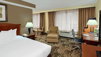 Habitación de hotel con una cama grande, dos sillones, un escritorio con lámpara y un ventanal con cortinas.