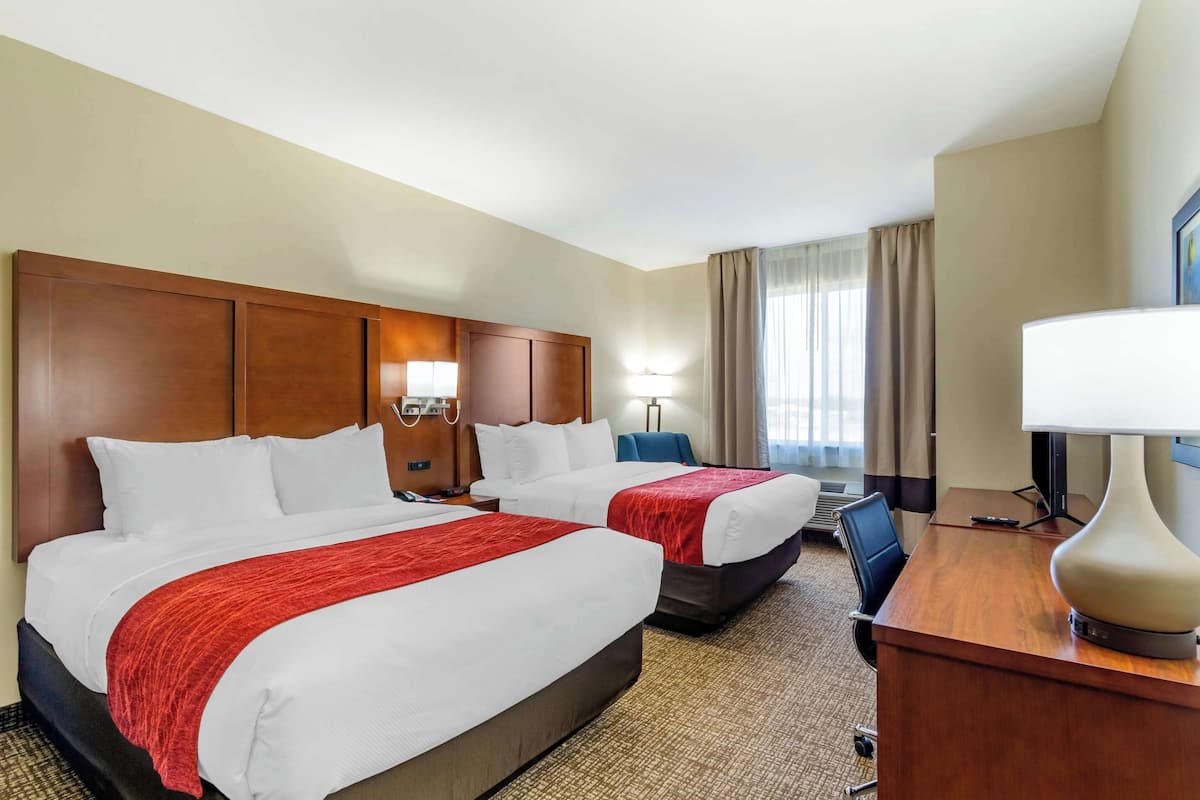 A hotel room with two beds, a desk, a lamp, and a view of the outdoors.