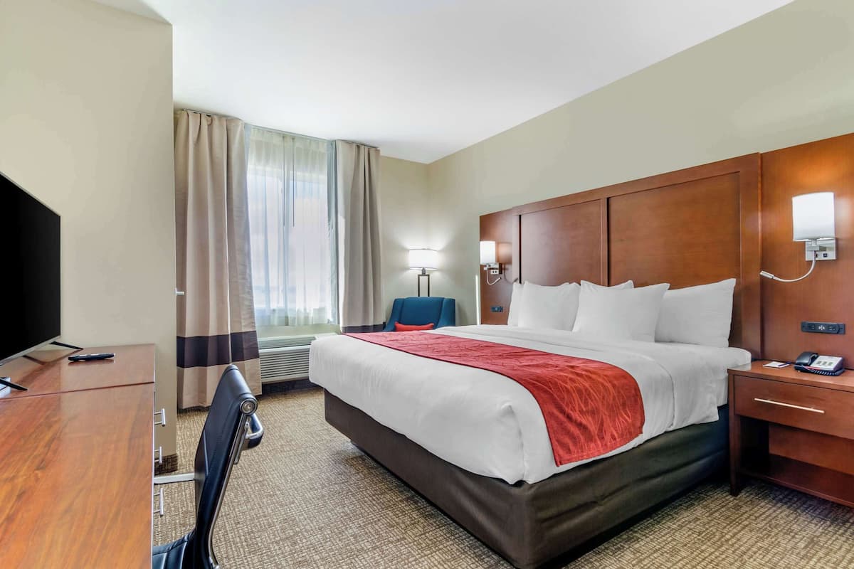 A hotel room with a large bed, a desk, a chair, a TV, a lamp, and a window with curtains.