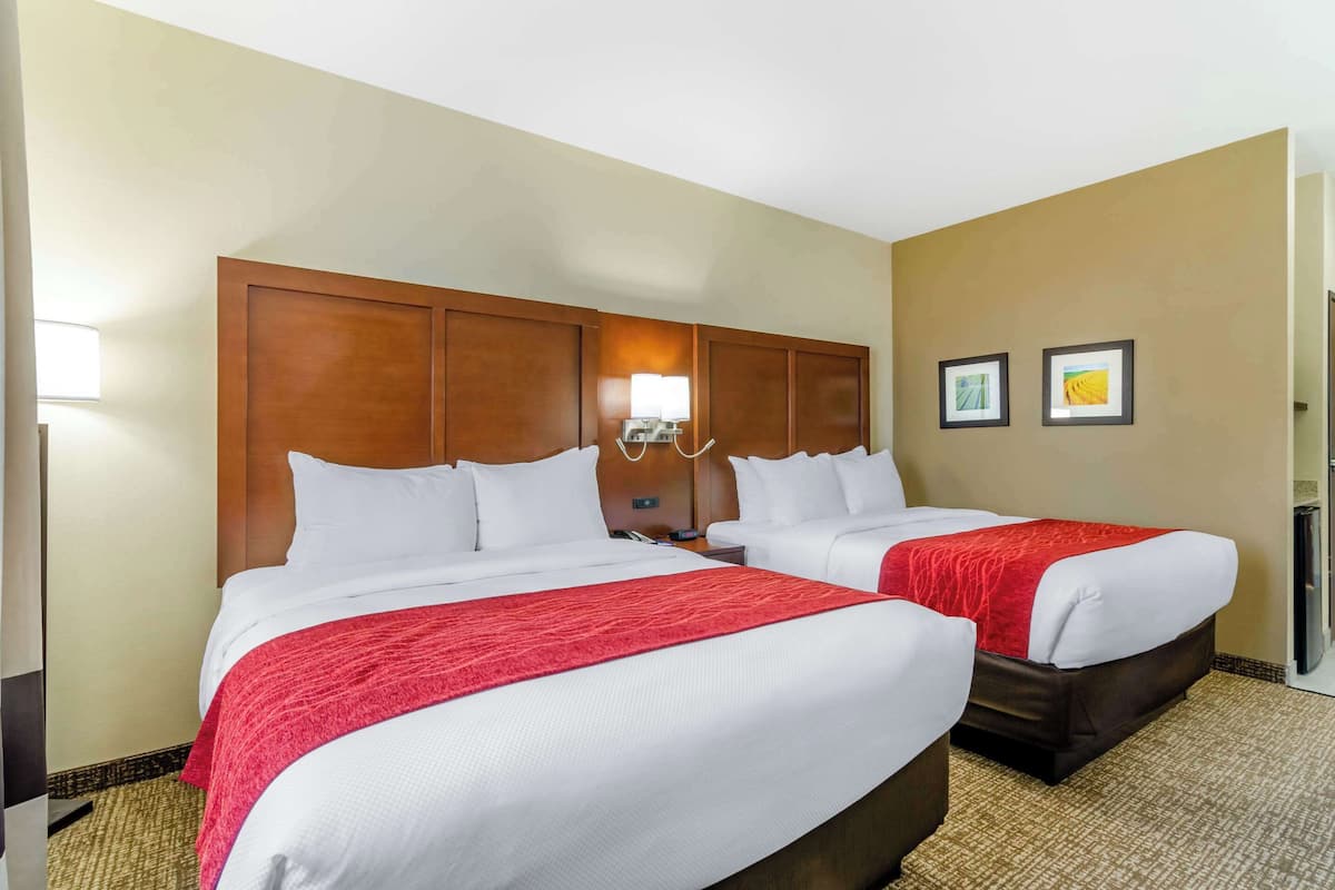 A hotel room with two beds, a wooden headboard, a bedside table with a lamp, and two framed pictures on the wall.