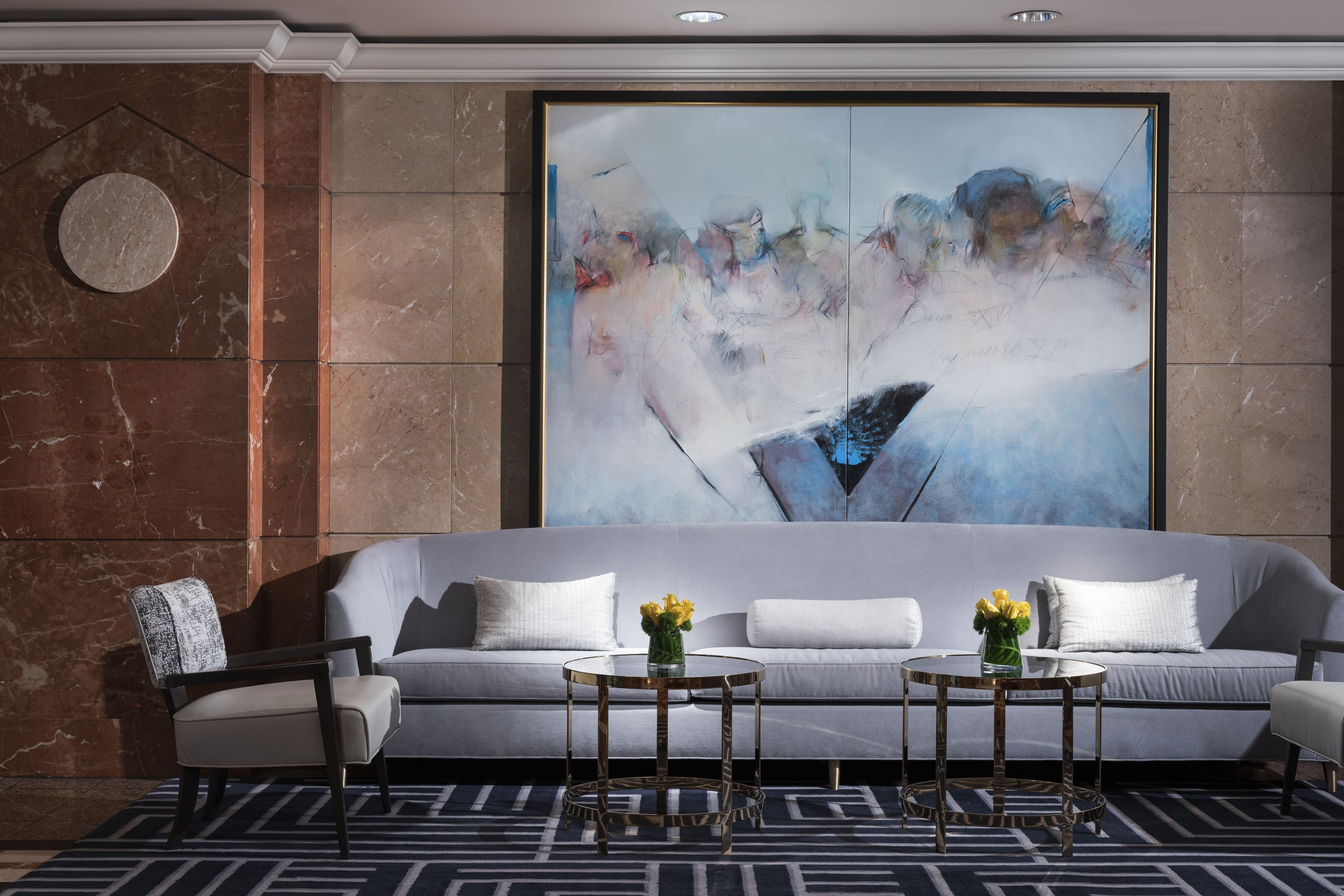 Een moderne lobby met een grijze bank, twee ronde tafels, een stoel en een groot abstract schilderij aan de muur.