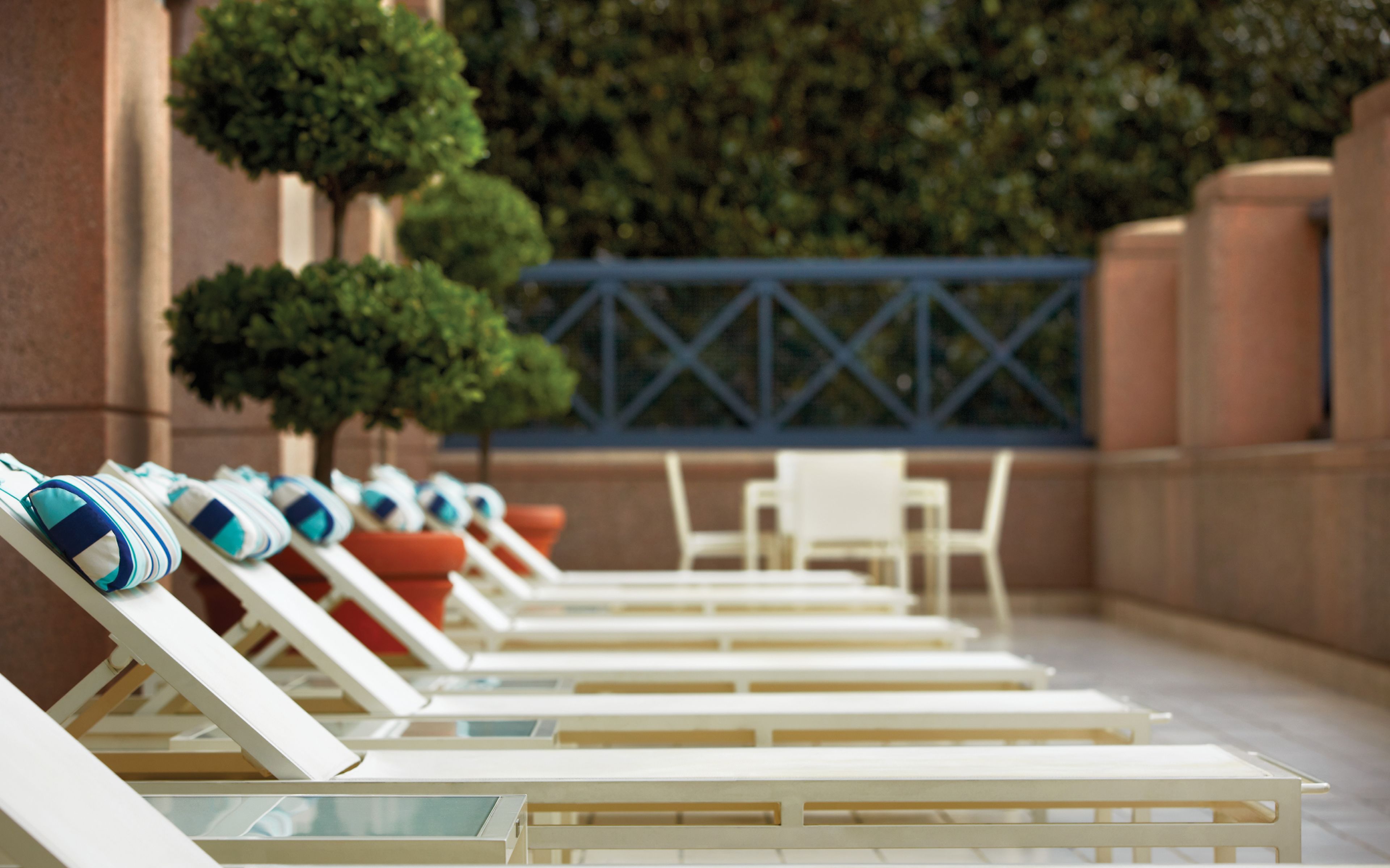 A row of sun loungers with towels, a small table, and chairs, set against a building facade with a blue railing and greenery.