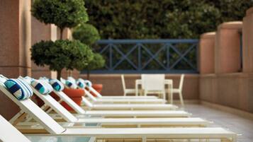A row of sun loungers with towels, a small table, and chairs, set against a building facade with a blue railing and greenery.