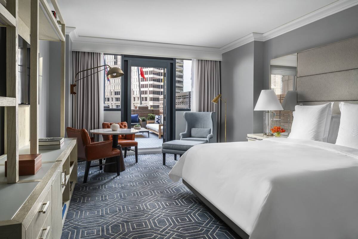 A hotel room with a large bed, a desk, a chair, a small table, and a view of the city through the window.