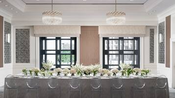 A banquet hall with a long table set for a formal event, featuring clear chairs and floral centerpieces.