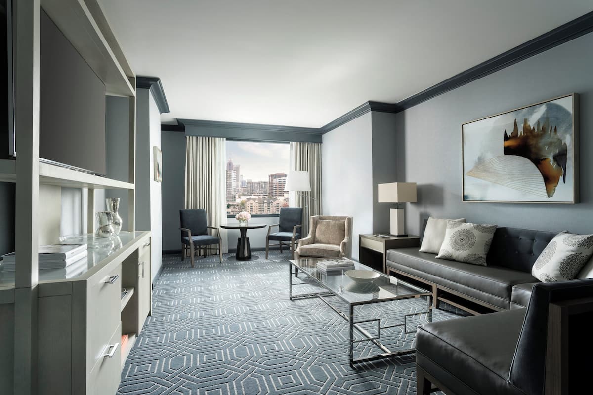 A modern hotel room with a city view, a sofa, a coffee table, and a dining area.