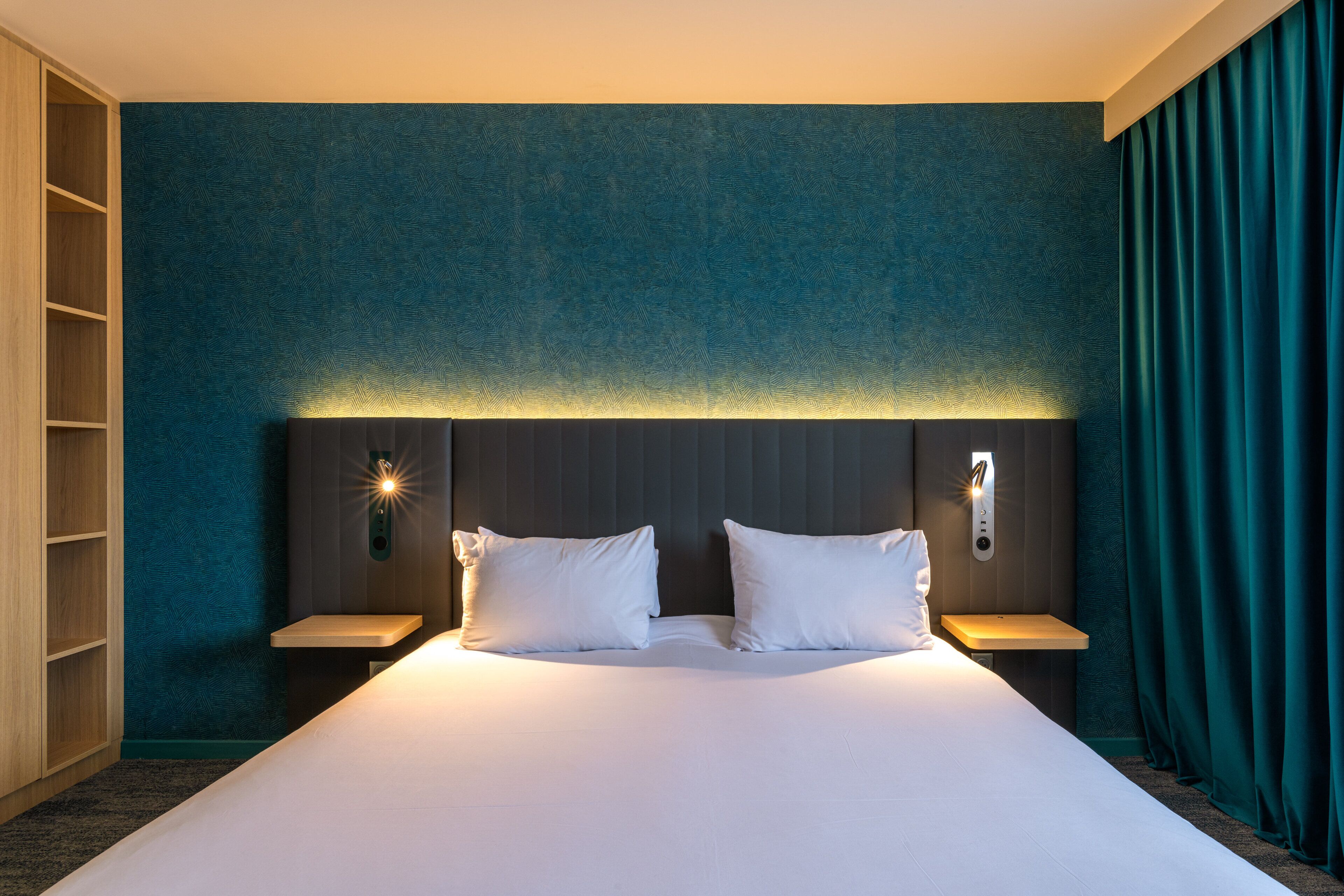Una cama grande con ropa de cama blanca, dos mesitas de noche y una lámpara fijada a la pared.
