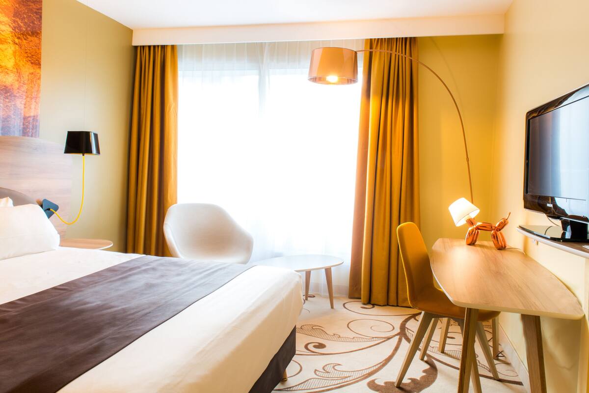 A hotel room with a bed, a desk with a lamp, a chair, a television, and a window with curtains.
