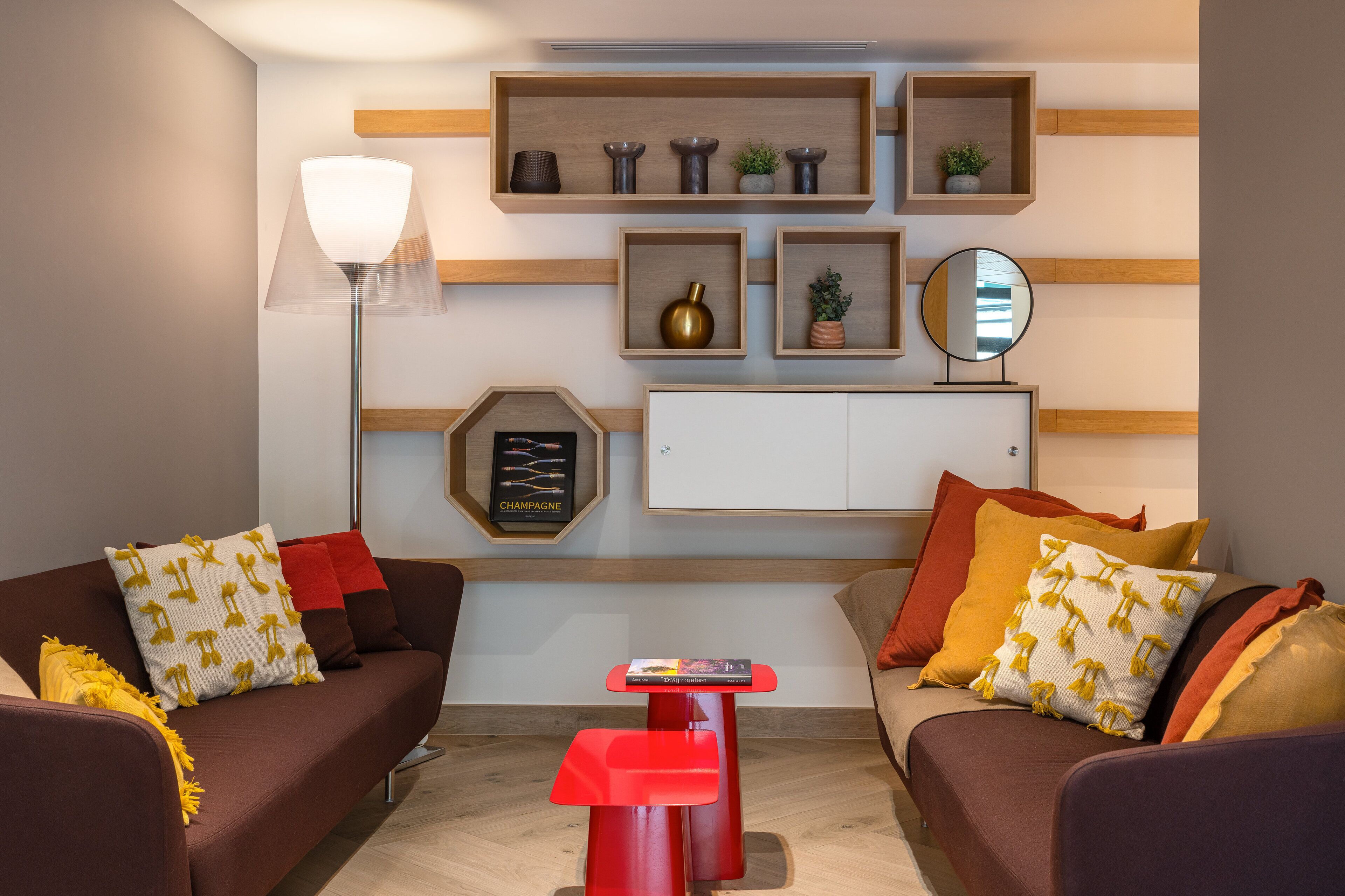 A modern living room with two sofas, a red side table, and a floor lamp. Shelves with decorative items and a framed picture are on the wall.
