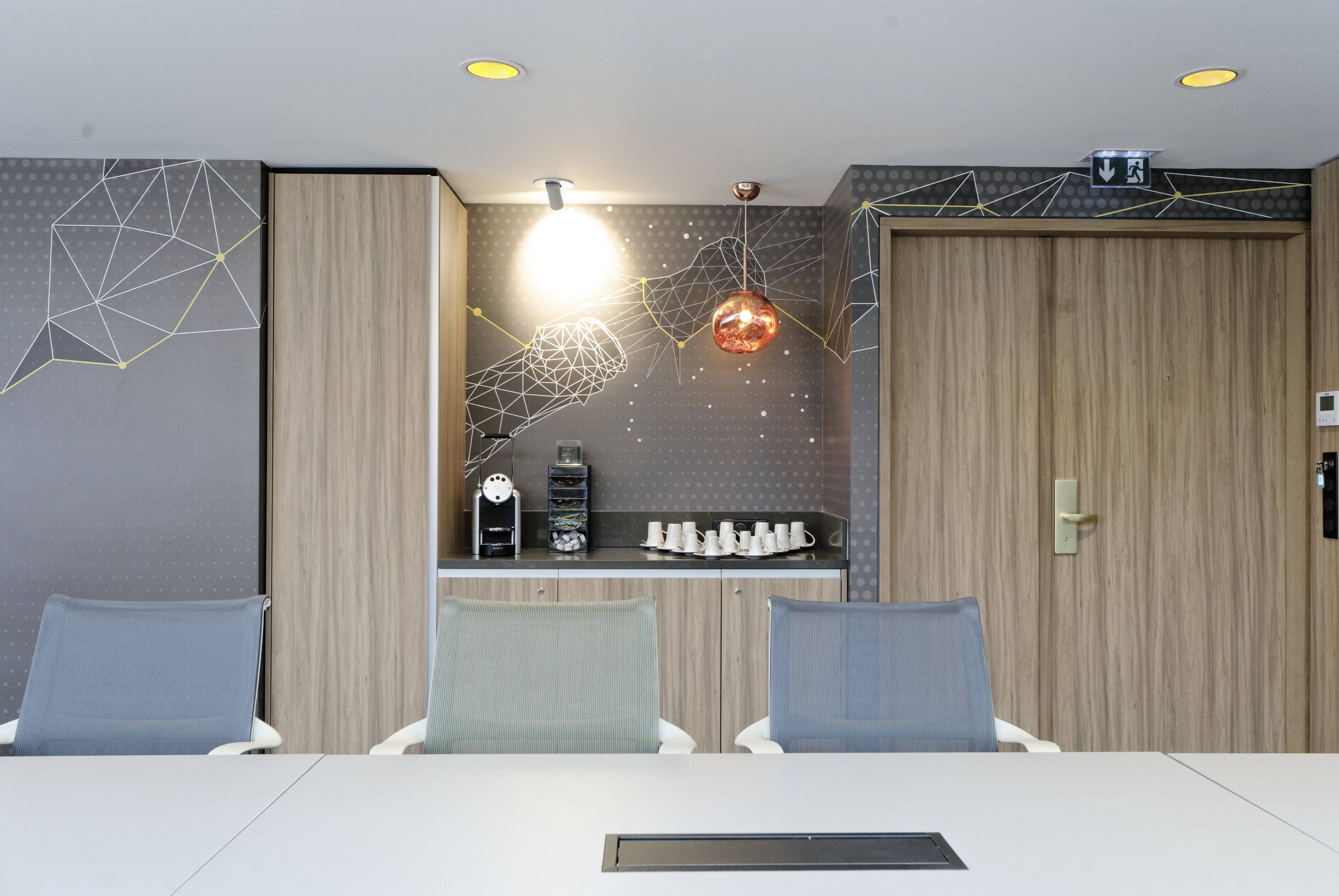 A conference room with a white table, two blue chairs, a coffee machine, and a wall with geometric patterns and a hanging light fixture.