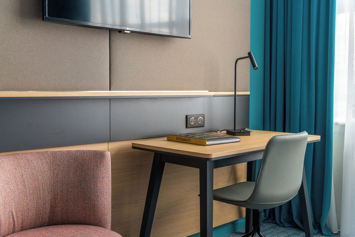 A hotel room with a wooden desk, a chair, a television mounted on the wall, and teal curtains.