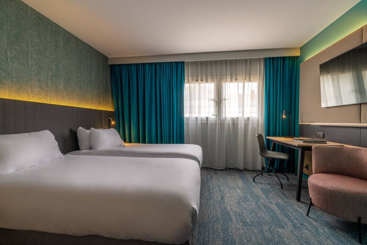 A hotel room with two beds, a desk, a chair, and a large window with curtains.