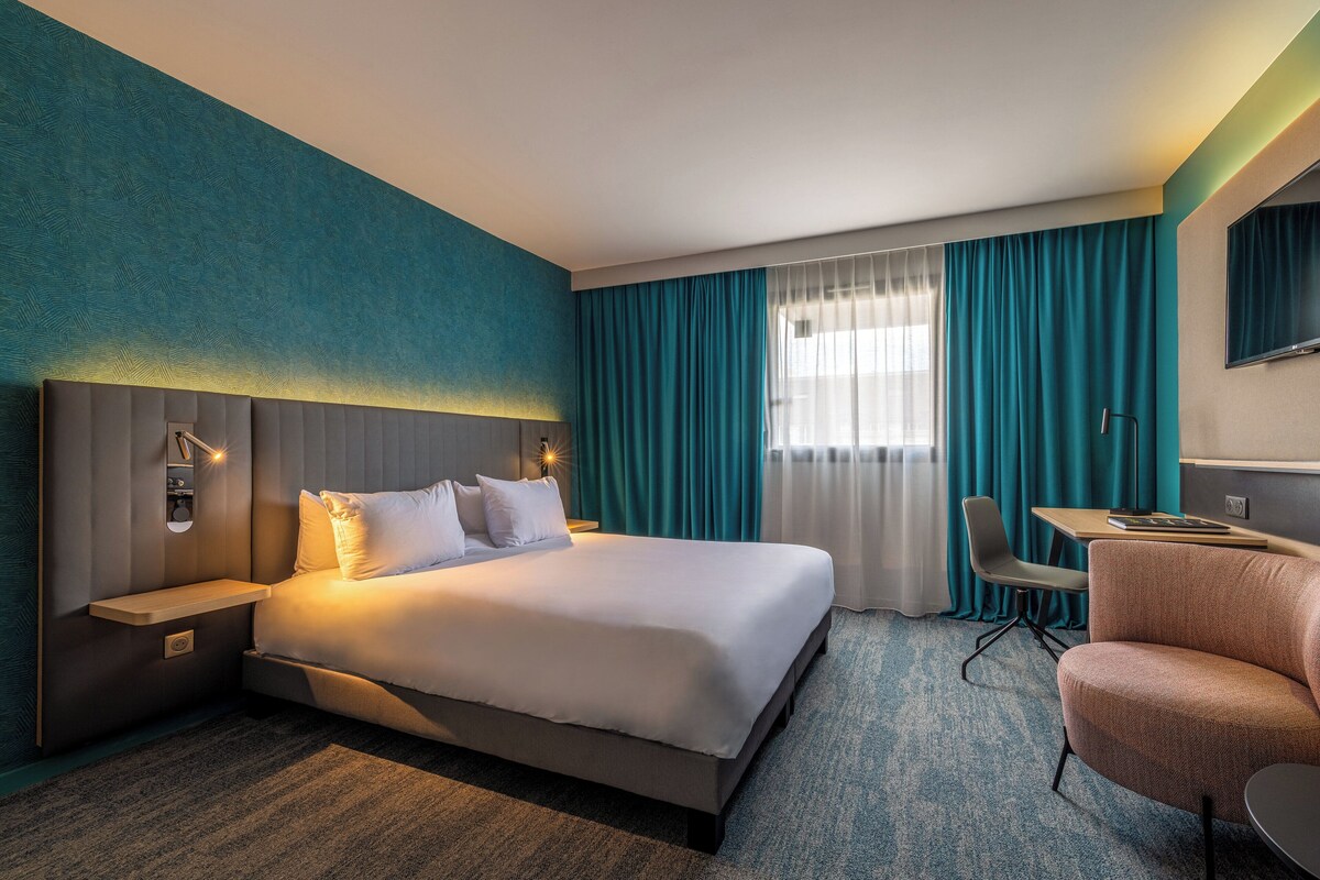 A hotel room with a large bed, a desk, a chair, and a TV.