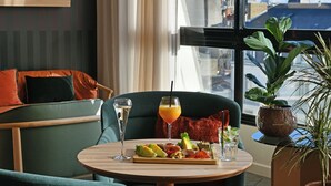 A modern hotel room with a dining area featuring a round table, green upholstered chairs, and a view of the city through large windows.