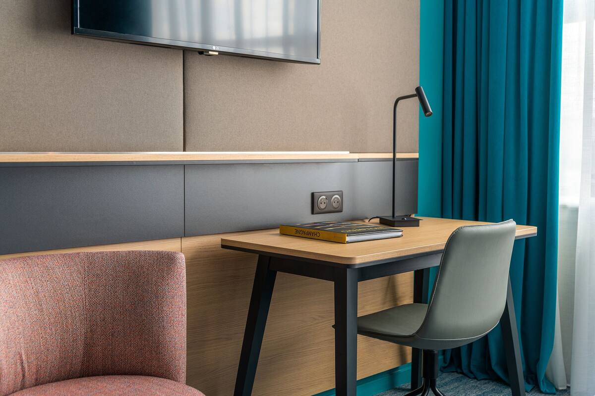 A hotel room with a wooden desk, a chair, a television mounted on the wall, and teal curtains.