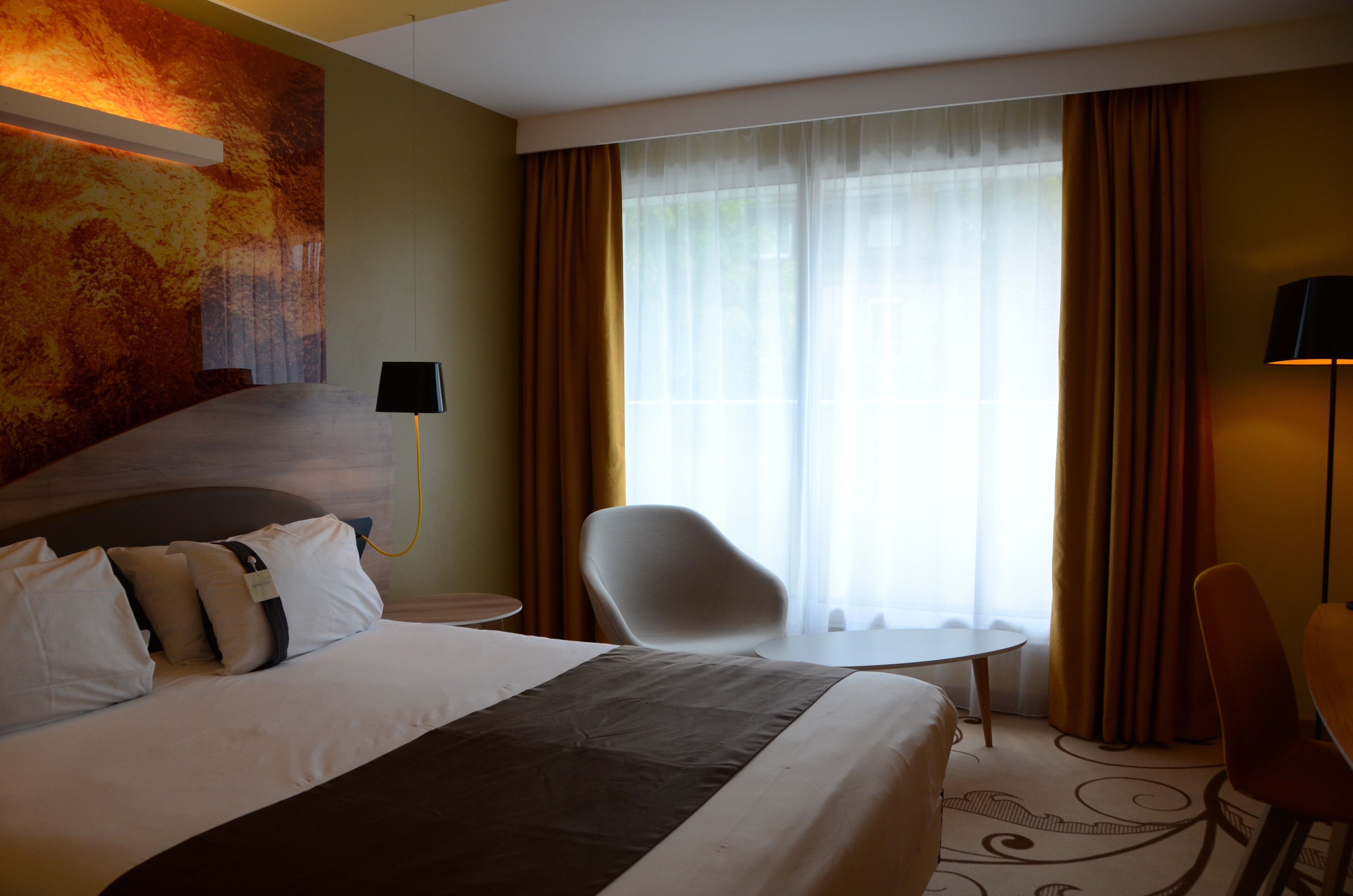 Una camera d'hotel con un letto grande, una scrivania, due sedie, una lampada e vista all'esterno attraverso tende leggere.