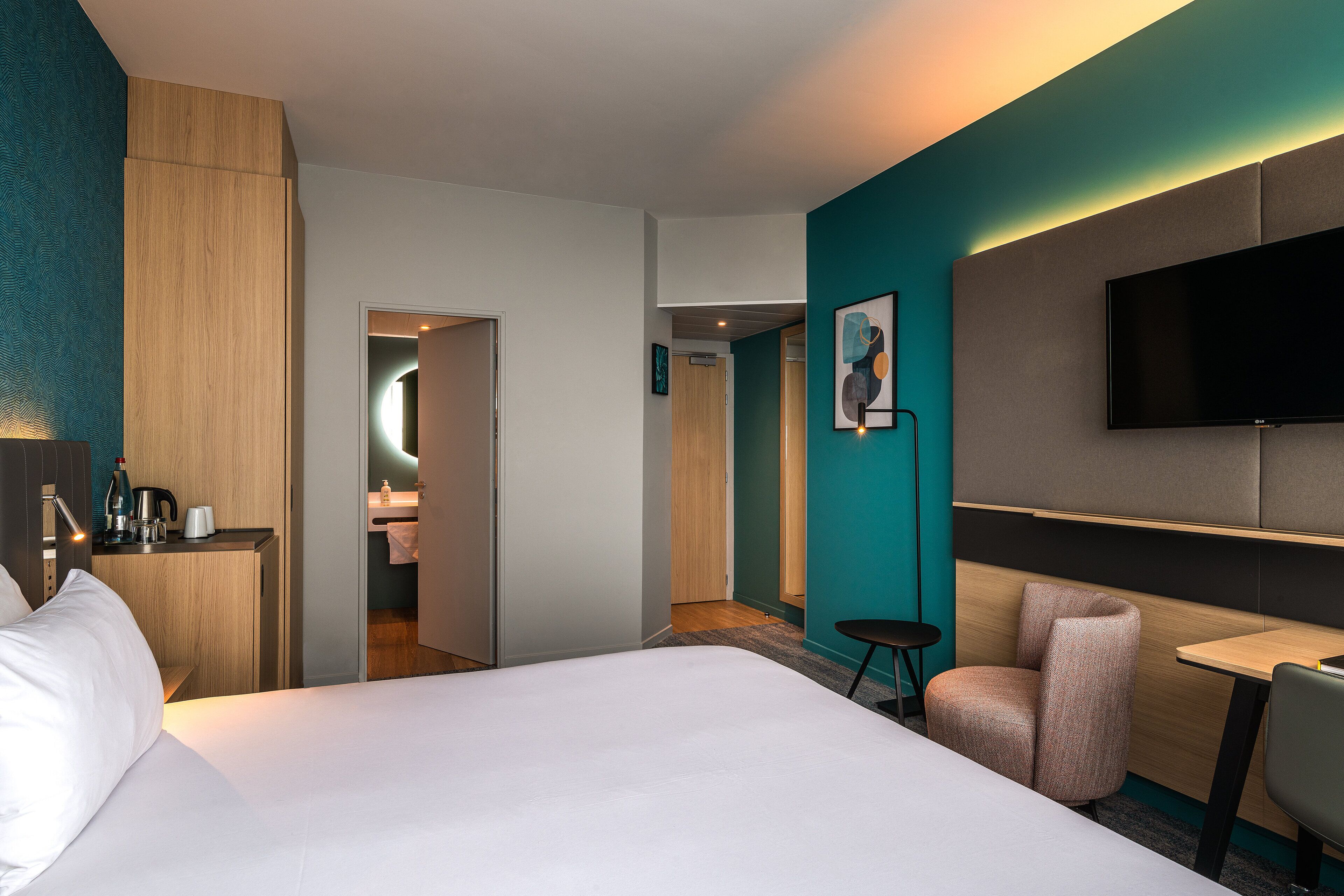 A hotel room with a bed, a desk, a chair, a TV, and a bathroom visible through an open door.