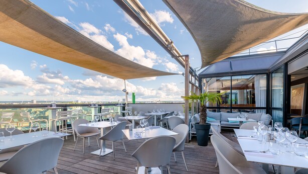 An outdoor dining area with tables and chairs, a view of the city, and a large umbrella providing shade.