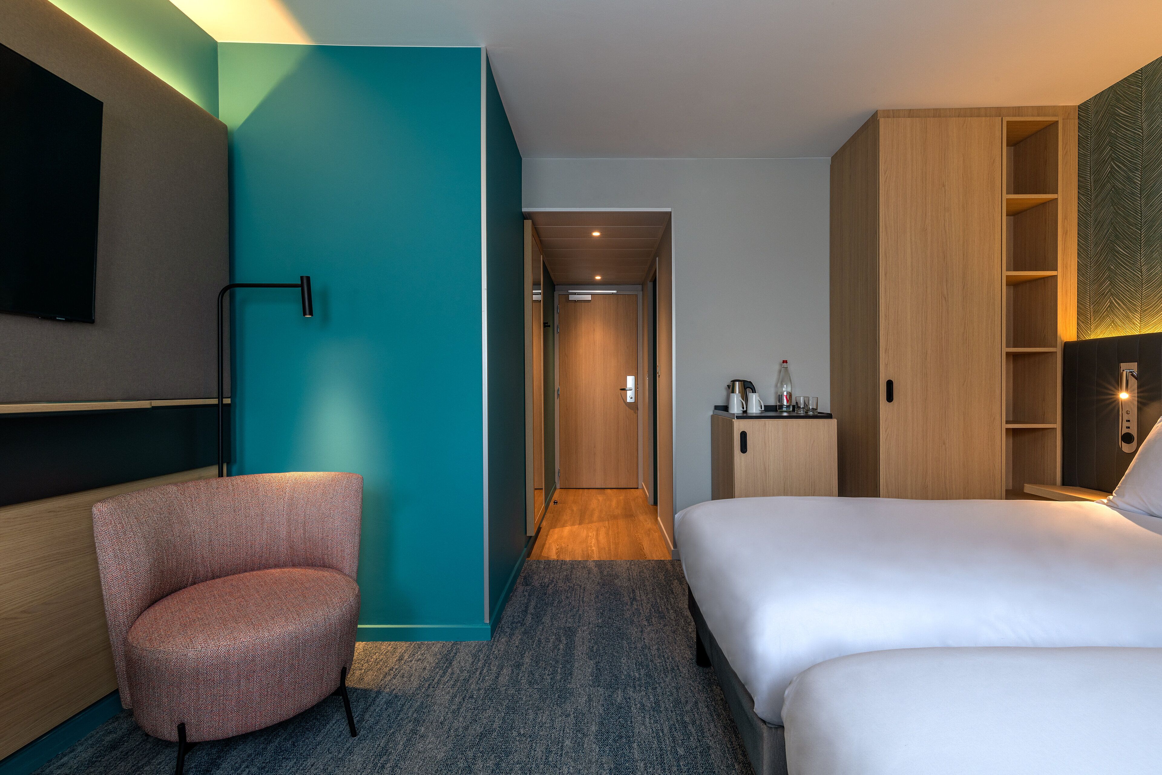 A modern hotel room with a large bed, a seating area, a TV mounted on the wall, and a wooden wardrobe.