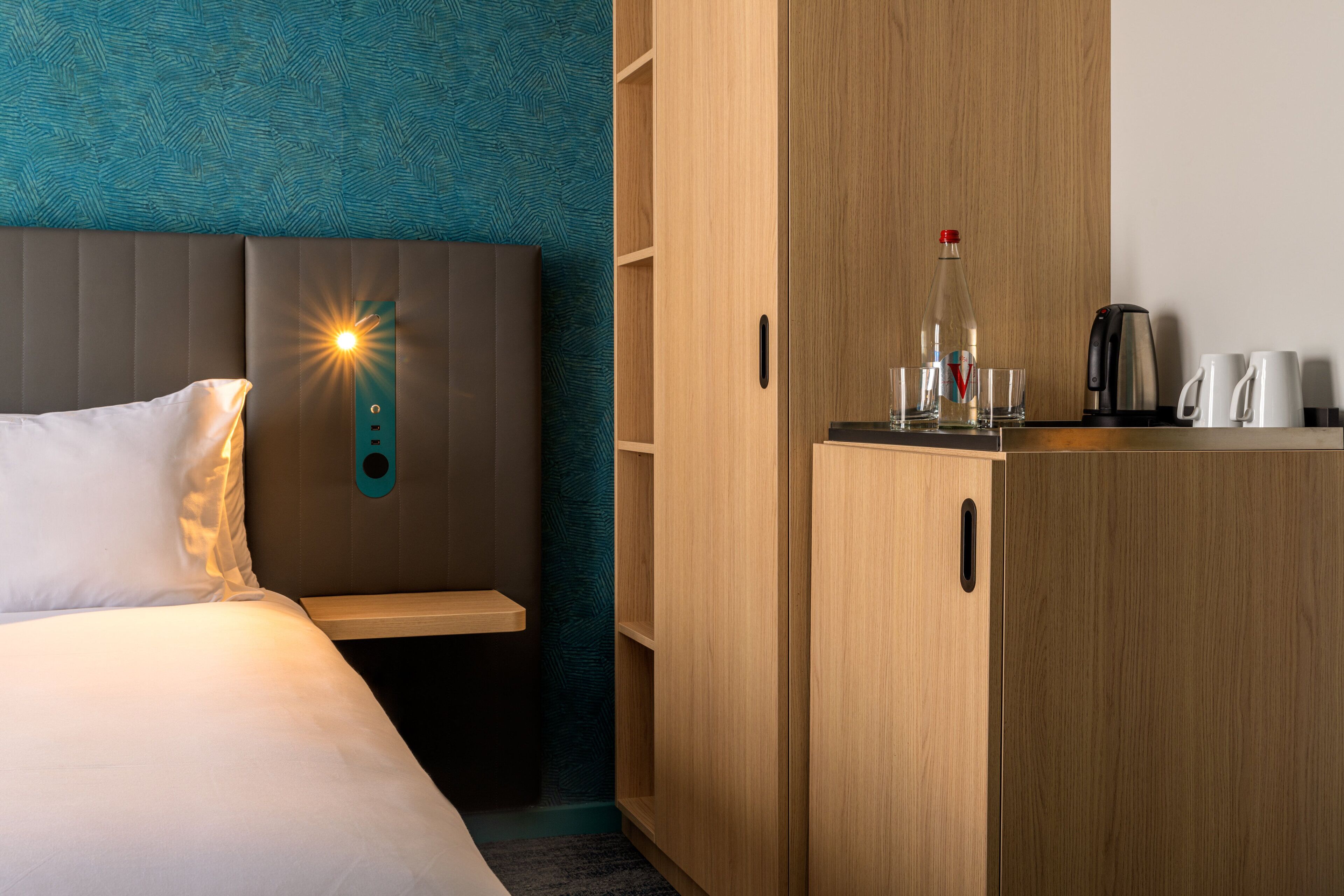 Habitación de hotel con cama, mesita de noche, armario de madera, cafetera y una botella de agua.