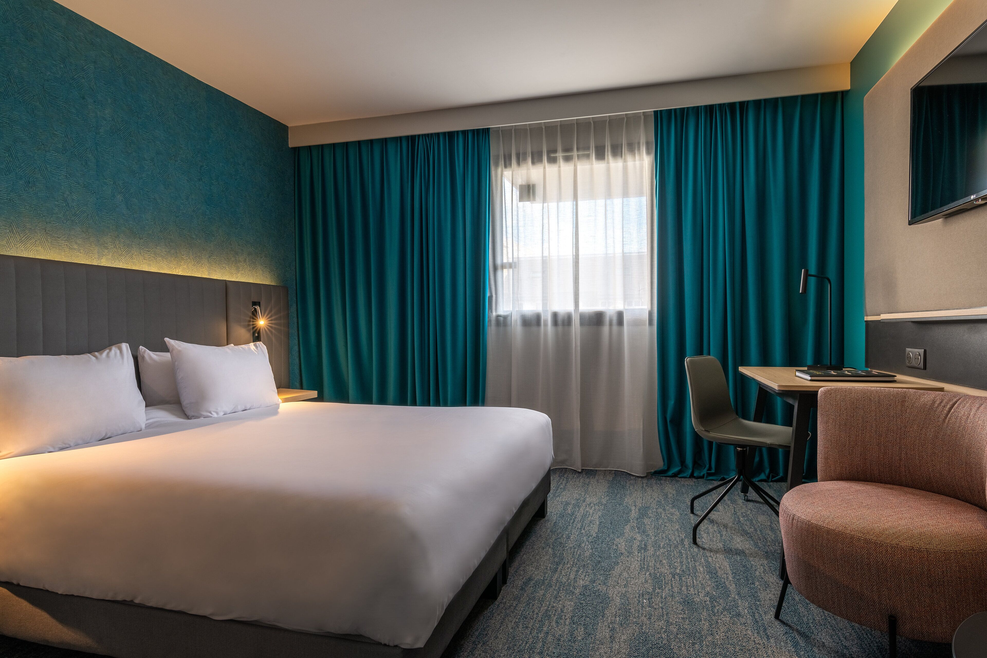 A hotel room with a large bed, a desk, a chair, and a TV on the wall.