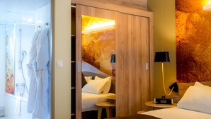 A hotel room with a bed, a bedside table with a lamp, a small stool, a shower area, and a robe hanging on a rack.