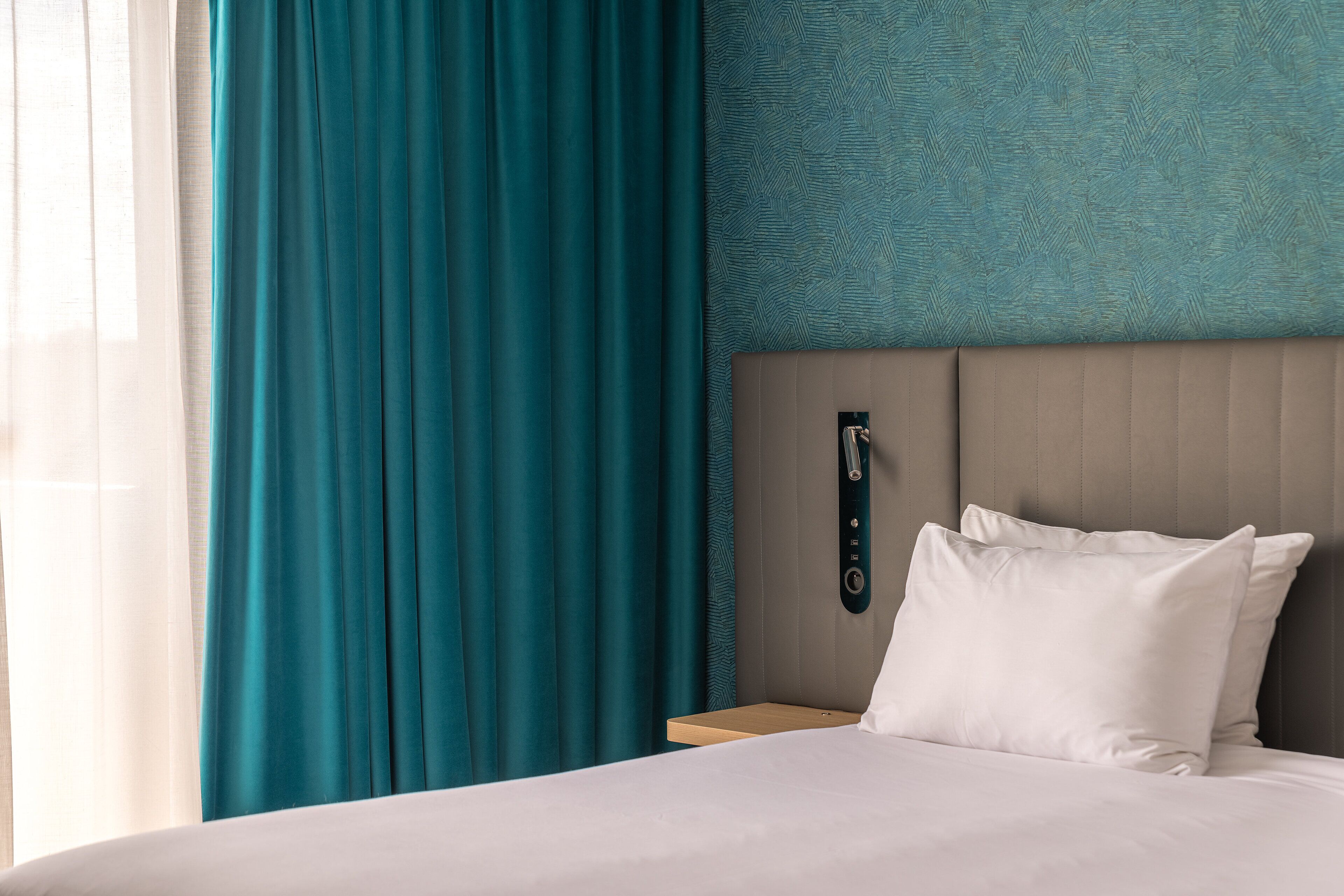 Een hotelkamer met een bed, blauwgroene gordijnen, een hoofdeinde met een bedieningspaneel en een behang met een patroon.