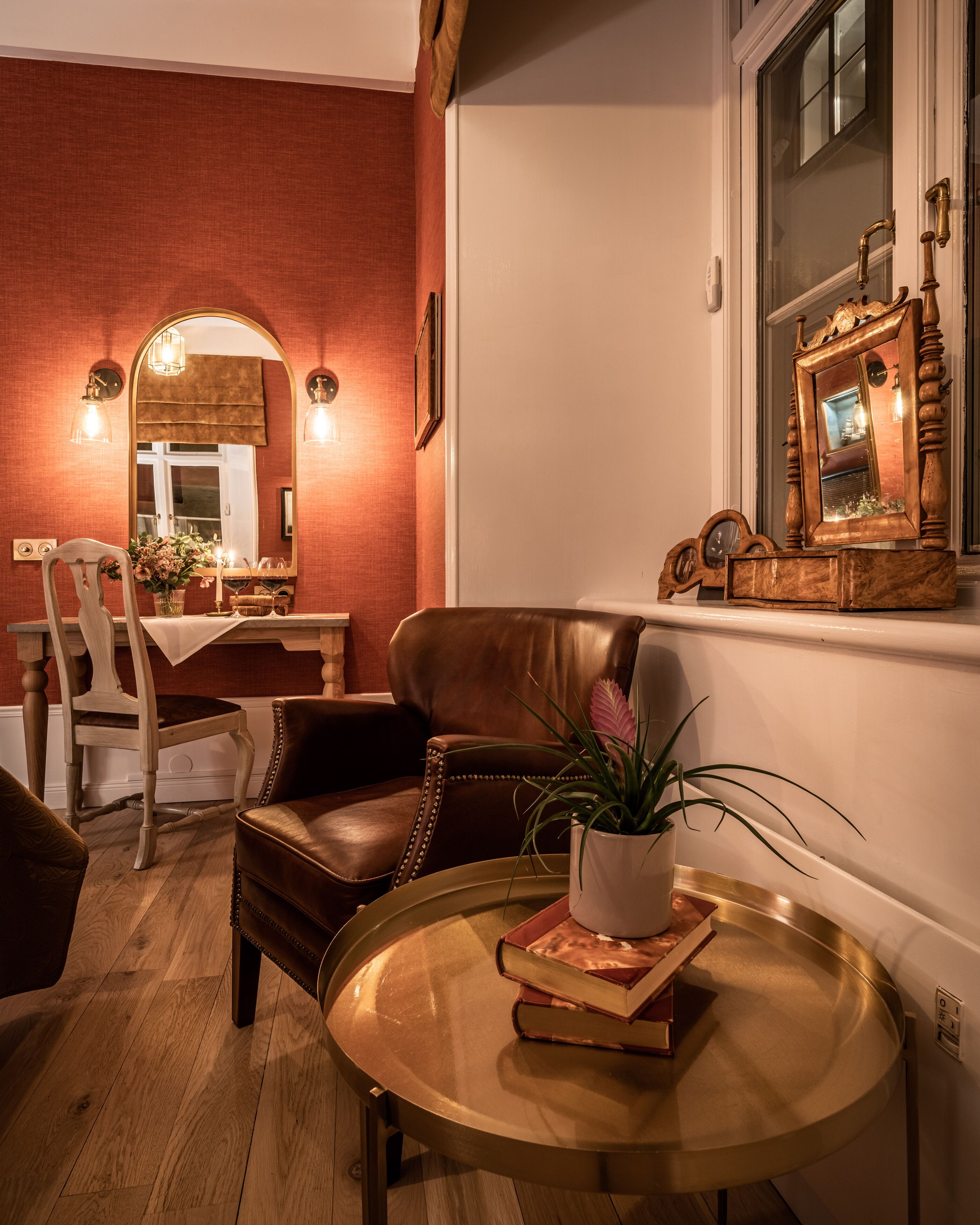 A cozy living room with a leather armchair, a round coffee table with books and a potted plant, a fireplace, and a large mirror with a decorative frame.